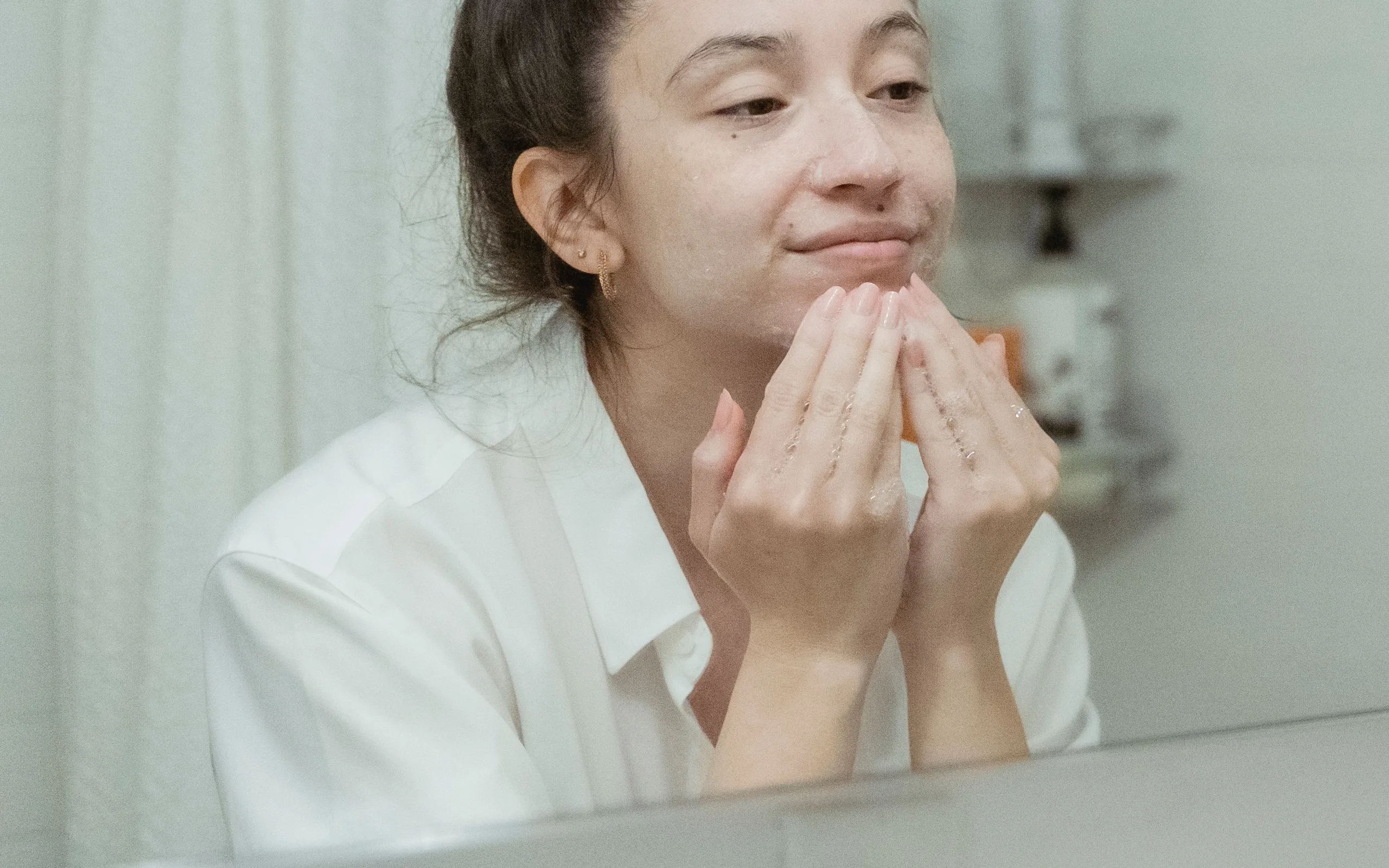 Femme appliquant un soin du visage devant un miroir de salle de bain : flacon de sérum posé sur le rebord et serviette propre à côté.